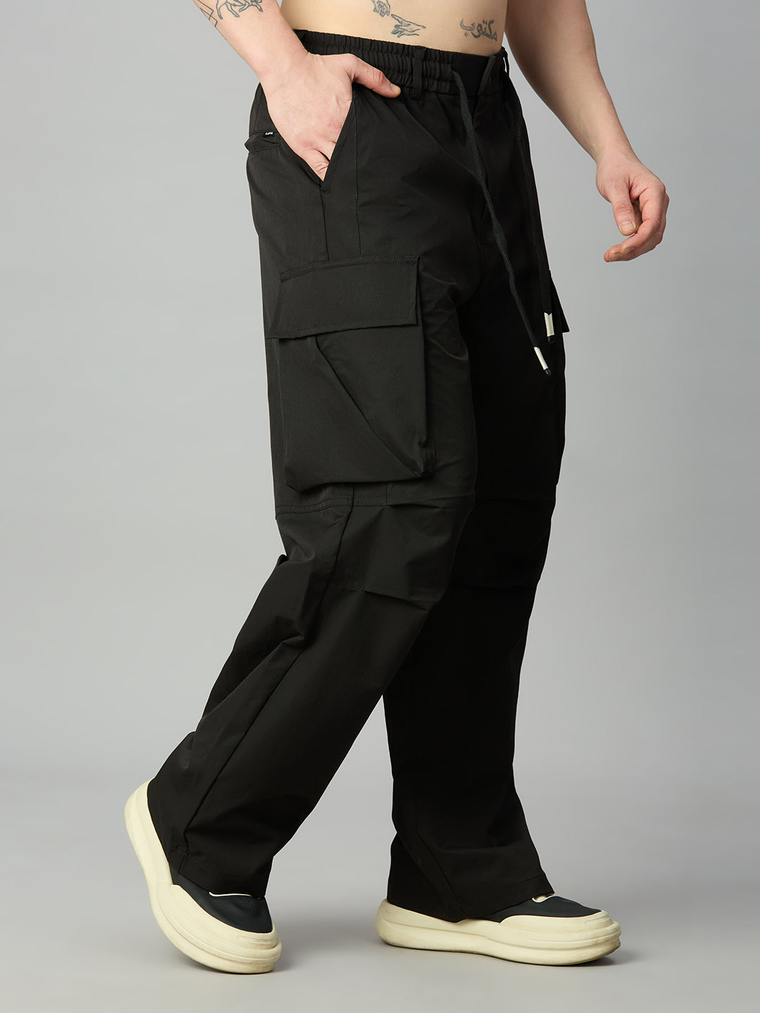 A person wearing loose black cargo pants with a drawstring waist, shown from the waist down against a light gray studio background. The pants have large flap cargo pockets and side pockets, paired with cream platform sneakers.