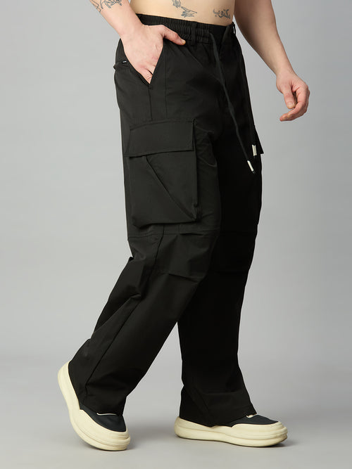 A person wearing loose black cargo pants with a drawstring waist, shown from the waist down against a light gray studio background. The pants have large flap cargo pockets and side pockets, paired with cream platform sneakers.