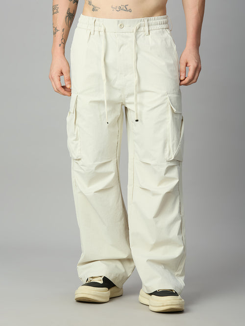 A person wearing very loose off-white cargo pants with a drawstring waist and large side pockets, paired with cream sneakers, photographed against a plain light gray studio background.
