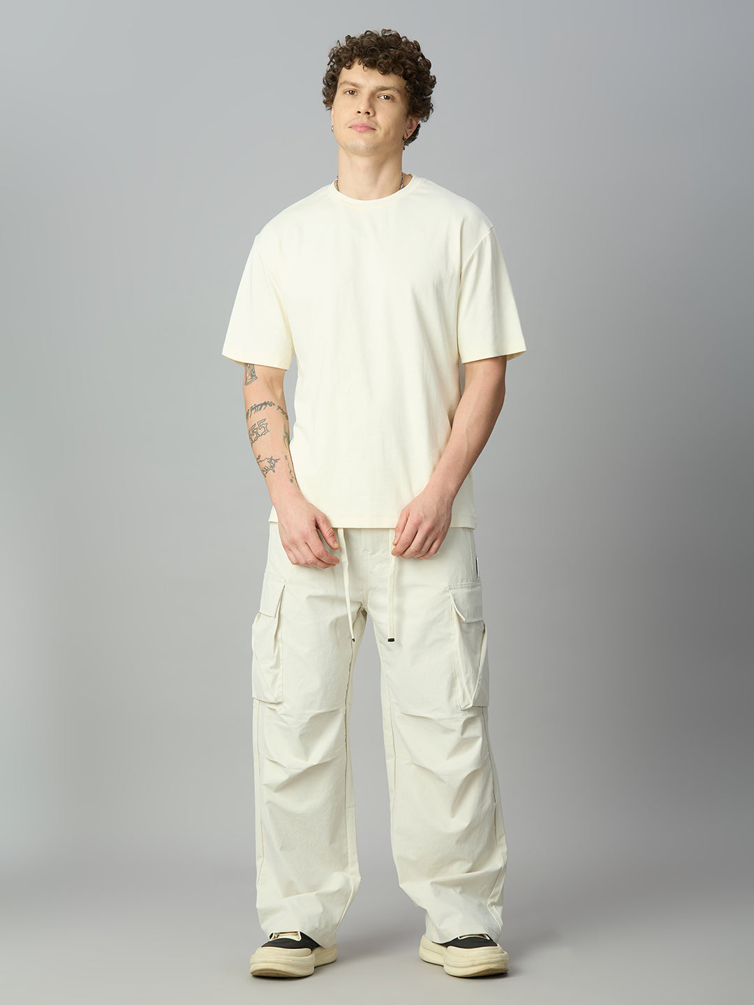 A man modeling a cream-colored short-sleeve t-shirt and matching cream cargo pants against a neutral gray studio background.