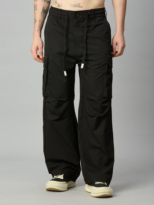 A person wearing black wide-leg cargo pants with a drawstring waist and large side cargo pockets, paired with cream sneakers, photographed against a plain light gray studio background.