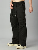 A person wearing loose black cargo pants with multiple flap and zip pockets, shown from the waist down against a light gray studio background. The pants are paired with black and cream sneakers.