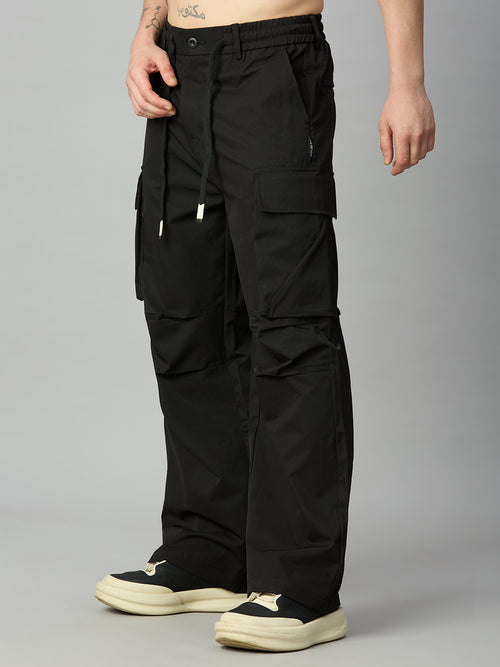 A person wearing loose black cargo pants with multiple flap and zip pockets, shown from the waist down against a light gray studio background. The pants are paired with black and cream sneakers.